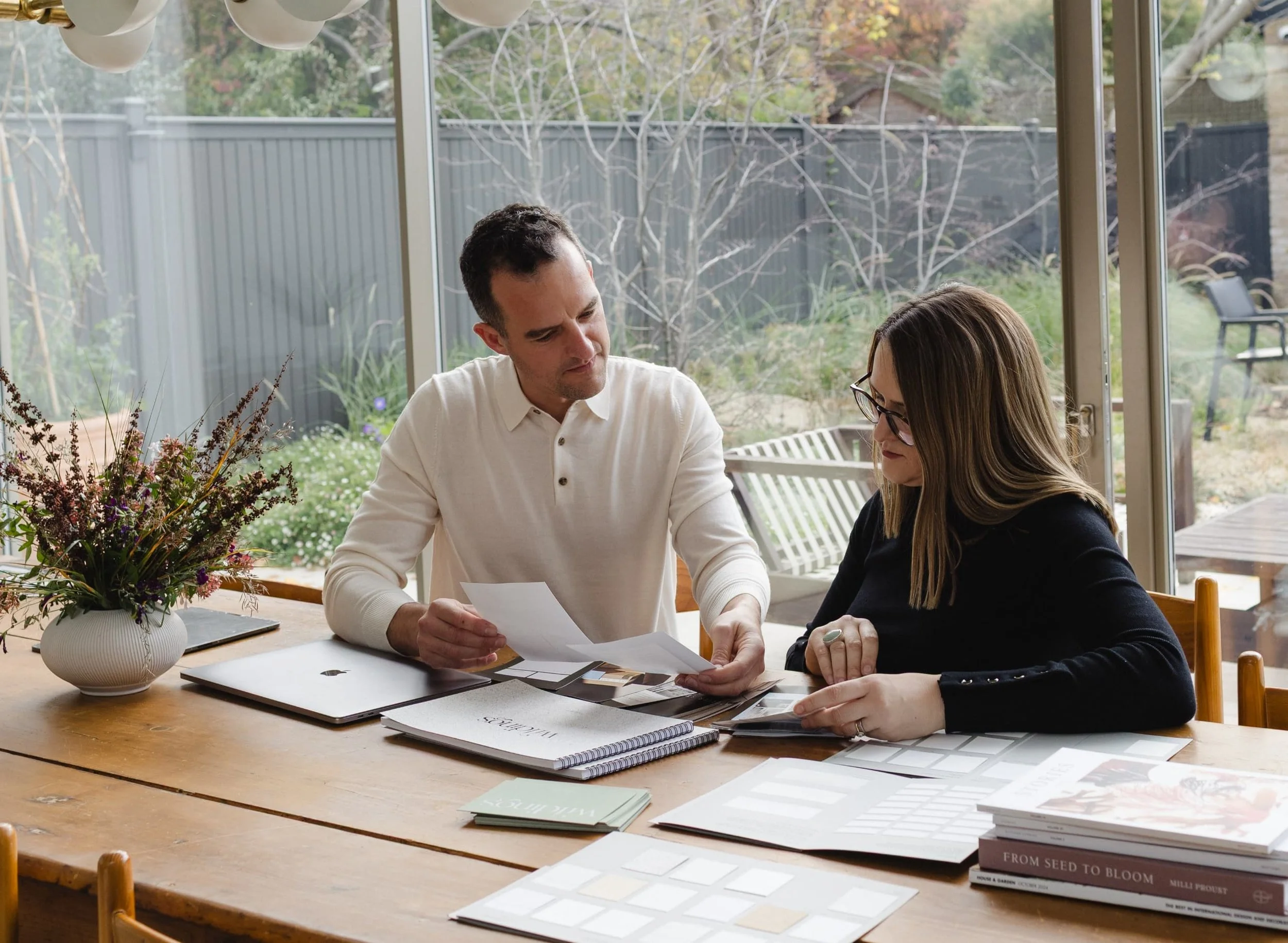 Creative duo reviewing documents at a large wooden table under a modern white light fixture by a glass wall