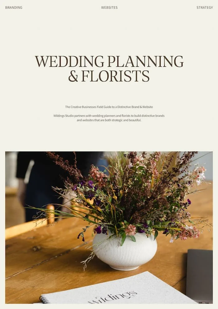 A white ribbed vase holding a colourful wild flower arrangement on a wooden table with a Wildings Studio notebook nearby