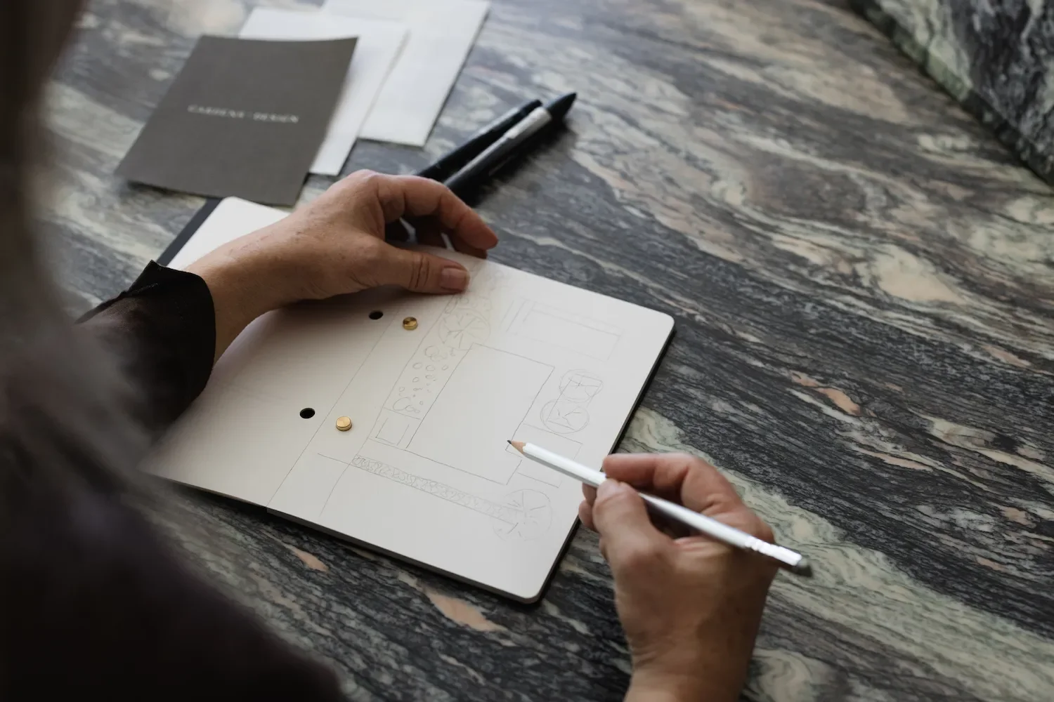 Close up of hands sketching a detailed garden site layout plan in a notebook alongside branded stationery on marble table