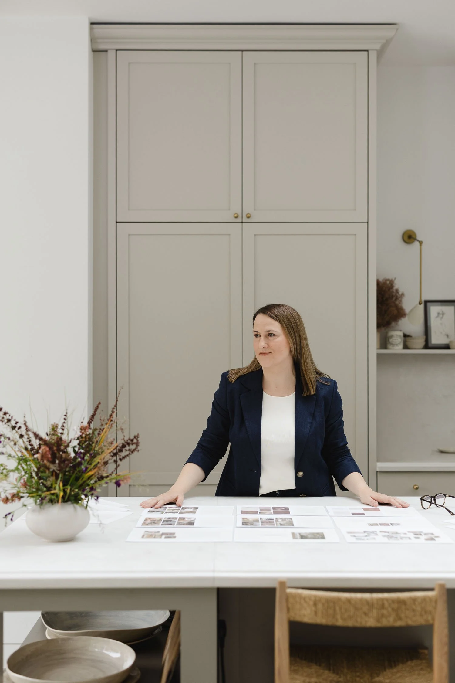 A female designer in a navy blazer standing at a white table reviewing various interior design project layouts and photos