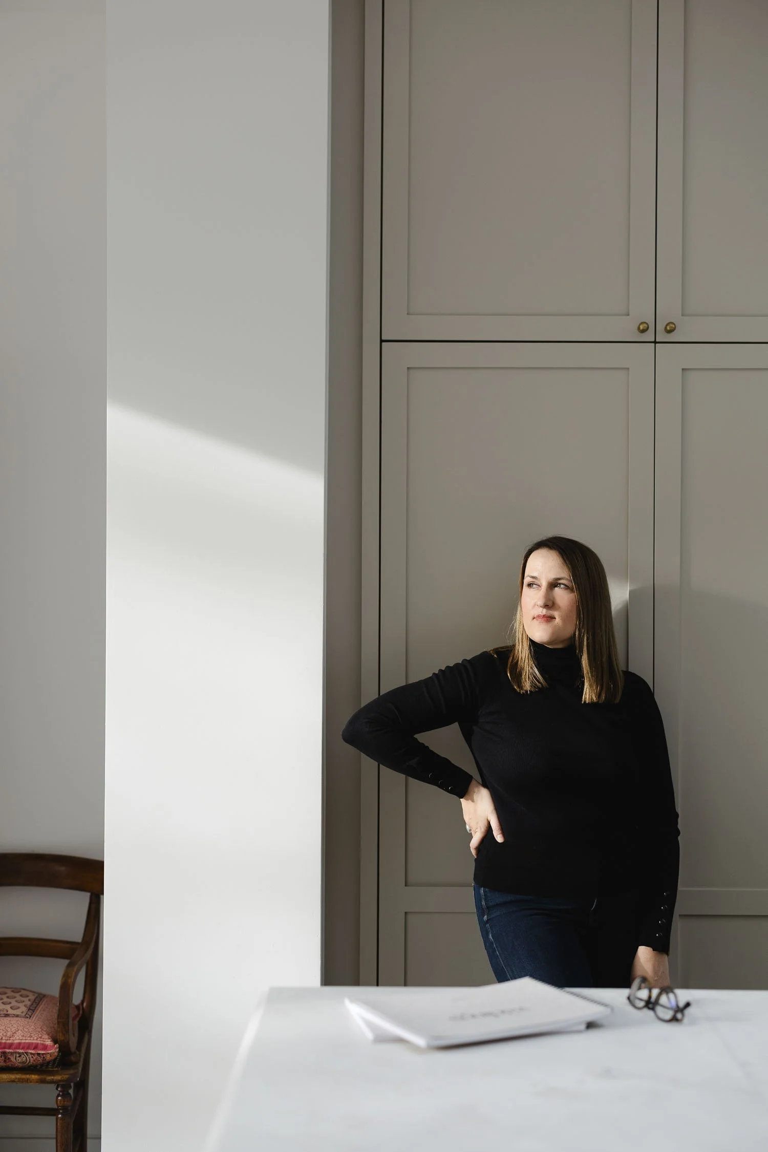 Woman in a black turtleneck and jeans standing by light grey cabinetry in a bright studio looking thoughtfully to the side