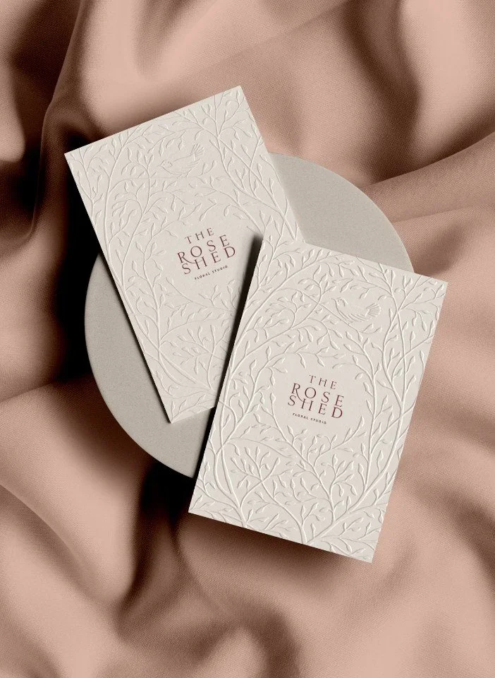 Two cream business cards for The Rose Shed floral studio with elegant embossed leaf patterns and bird motifs on pink fabric