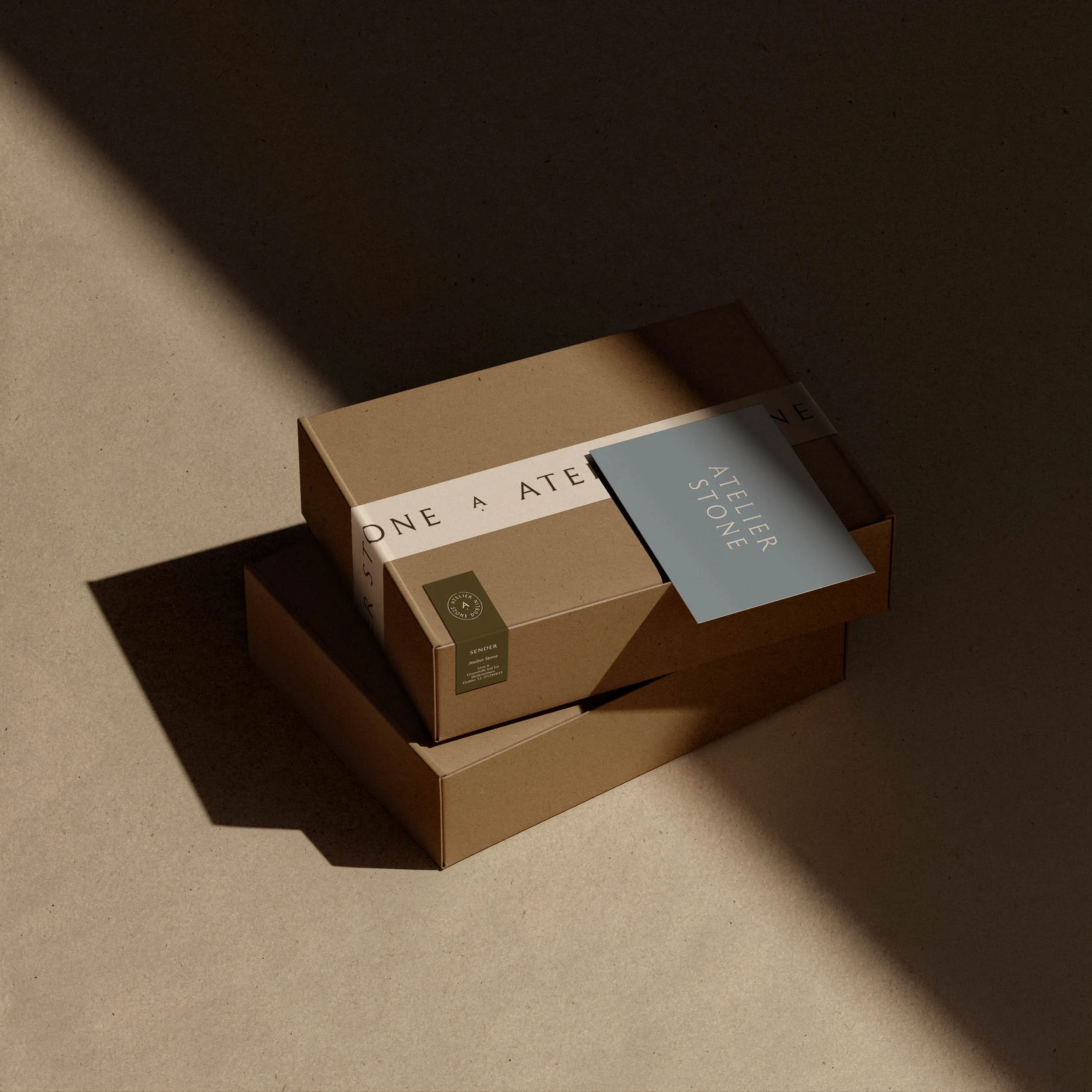 Two cardboard postal boxes with Atelier Stone branding tape and a blue brand card set against a minimal concrete background
