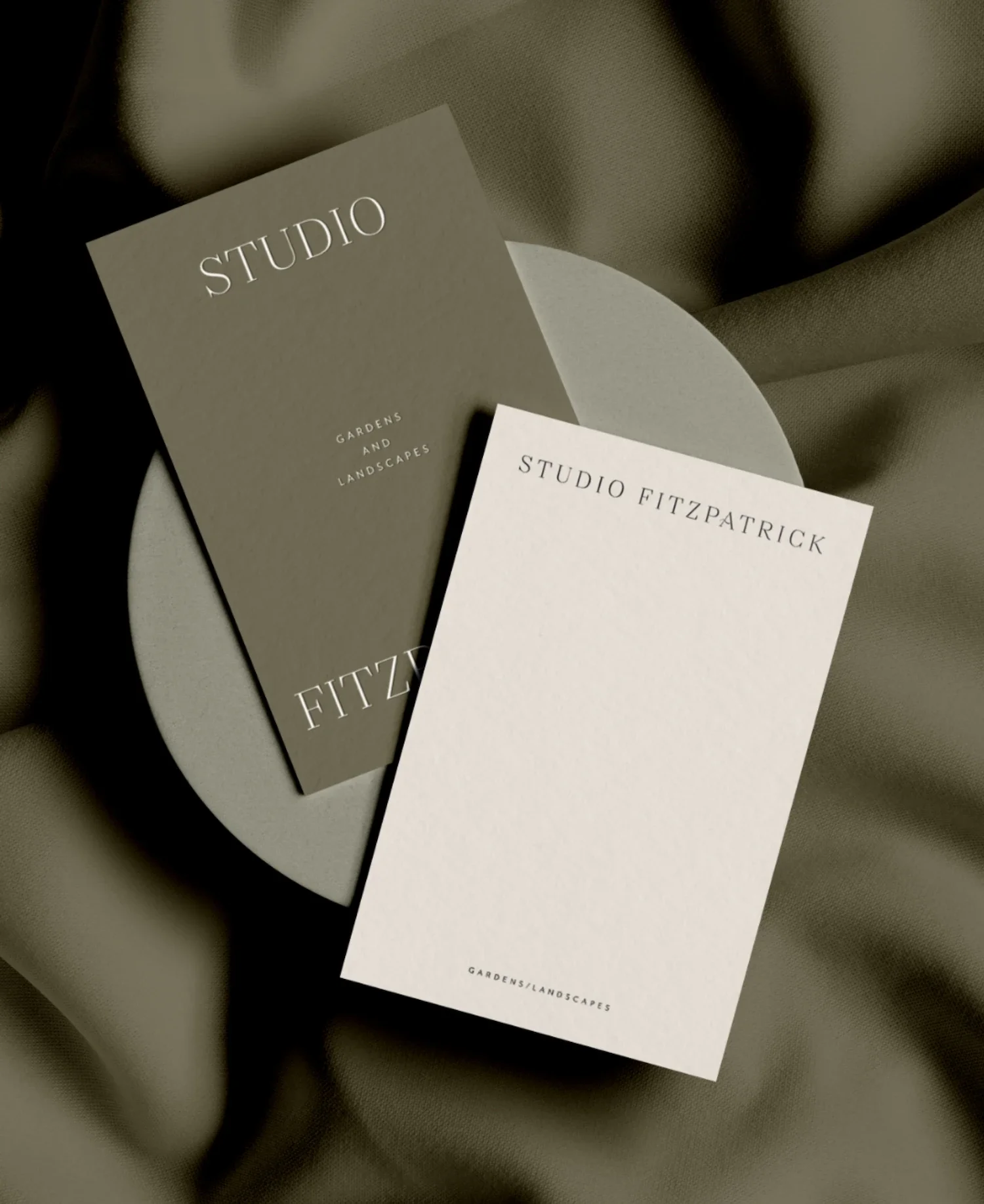 Premium olive green and cream business cards displayed on a contemporary ceramic disc against flowing silk fabric