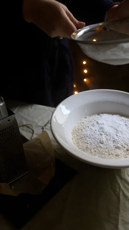 Hands using a metal sieve to sift white flour into a large ceramic bowl for making festive orange pastry dough