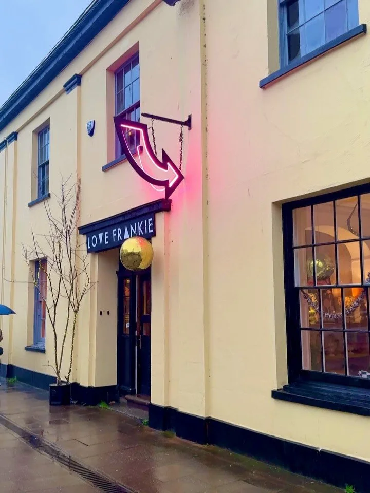 Exterior of Love Frankie shop in Totnes featuring a pale yellow facade with a neon arrow sign and a gold disco ball