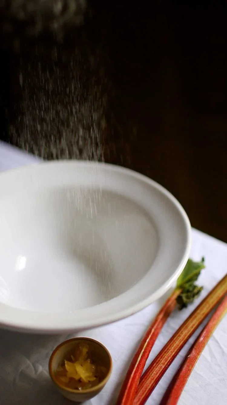 Flour being sifted into a large white ceramic bowl with fresh rhubarb stalks and ginger pieces visible in the foreground