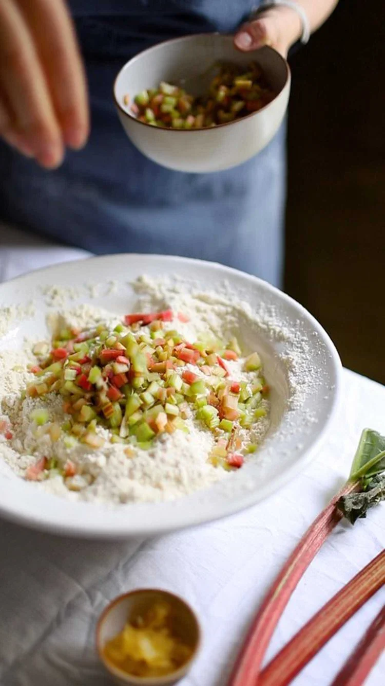 Hand sprinkling a bowl of freshly diced pink and green rhubarb over a flour base to prepare fruit scone dough