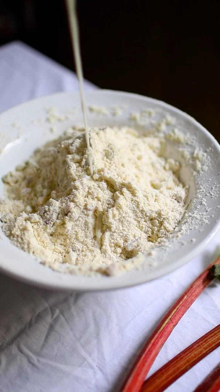 Milk being poured into a well in a bowl of dry ingredients to make scones with rhubarb stalks on a white cloth