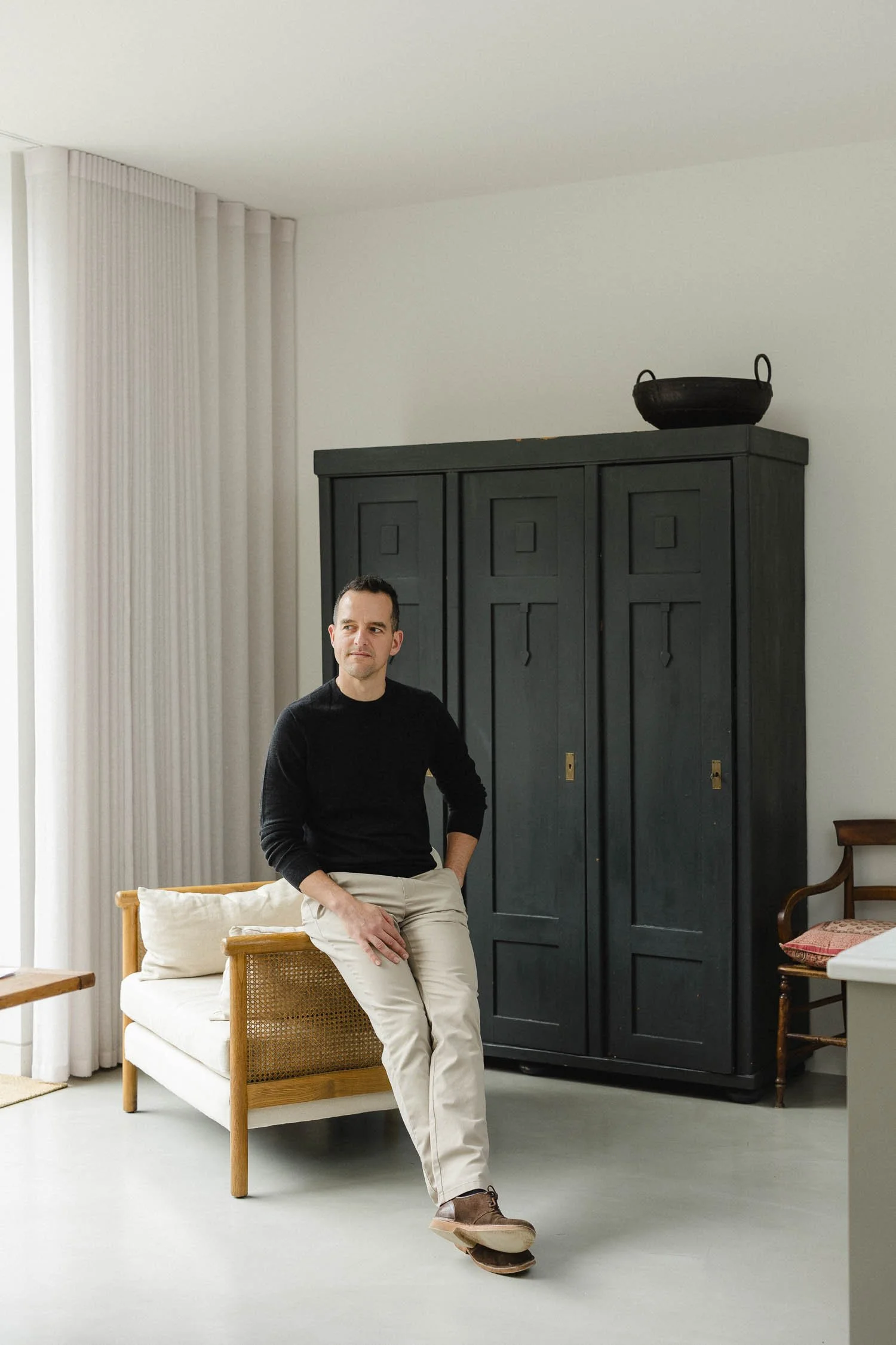 Designer in a black jumper sitting on a cane chair in front of a tall black vintage cupboard in a light room