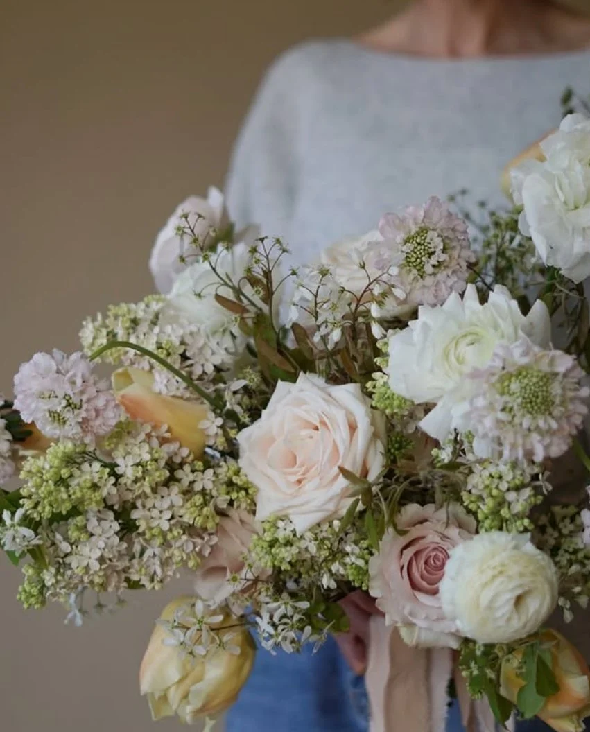 Person holding a large wedding bouquet featuring white ranunculus, pale roses, lilac and delicate green foliage