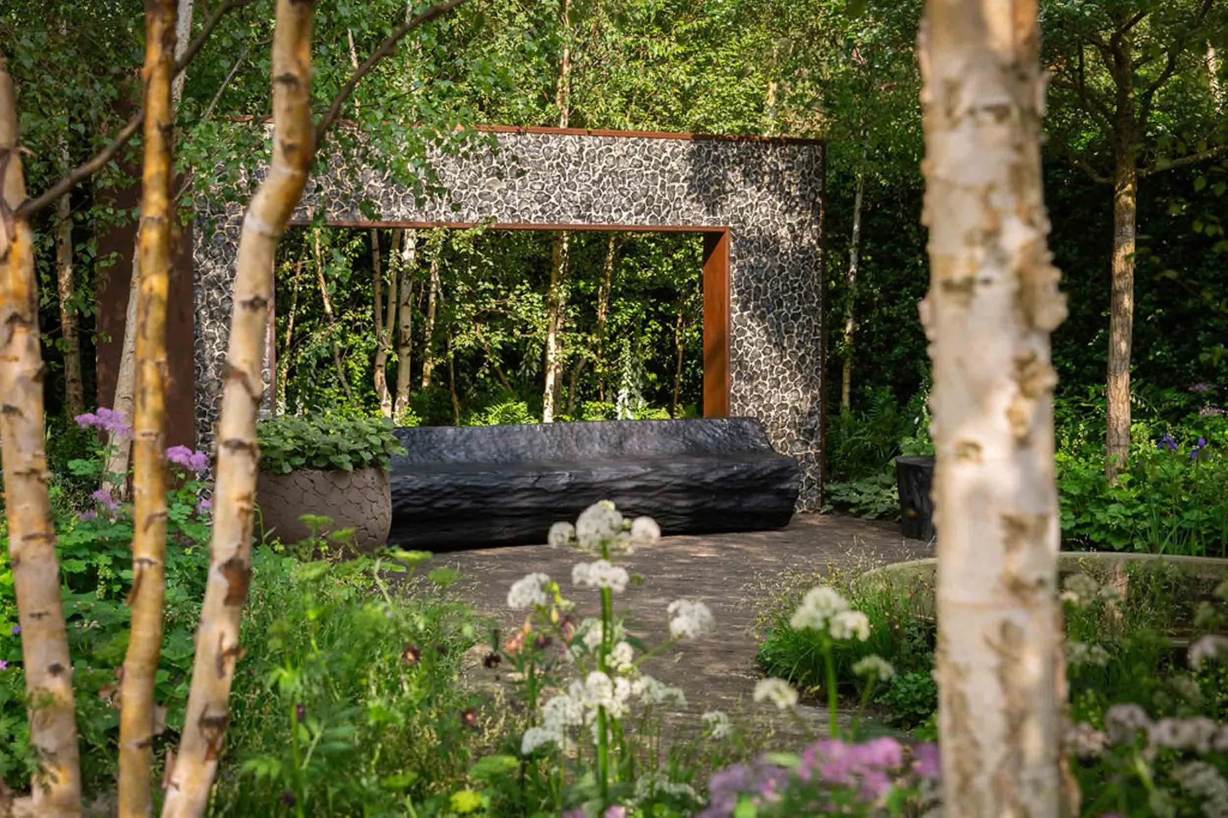 Charred black timber bench positioned in front of a stone textured wall frame surrounded by silver birch trees and ferns
