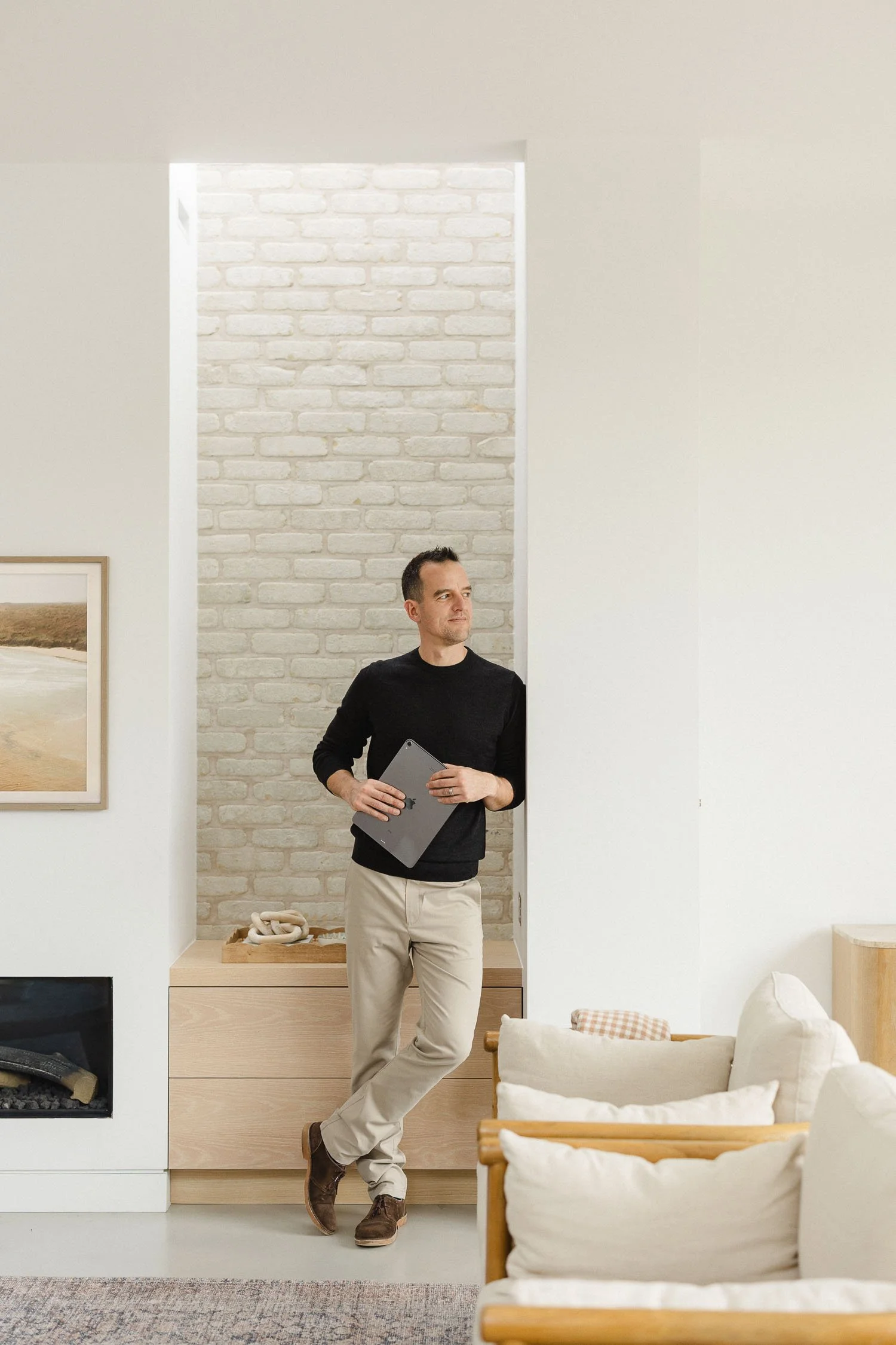 Man in a black jumper and beige trousers leaning against a white brick alcove while holding a grey tablet in a modern room