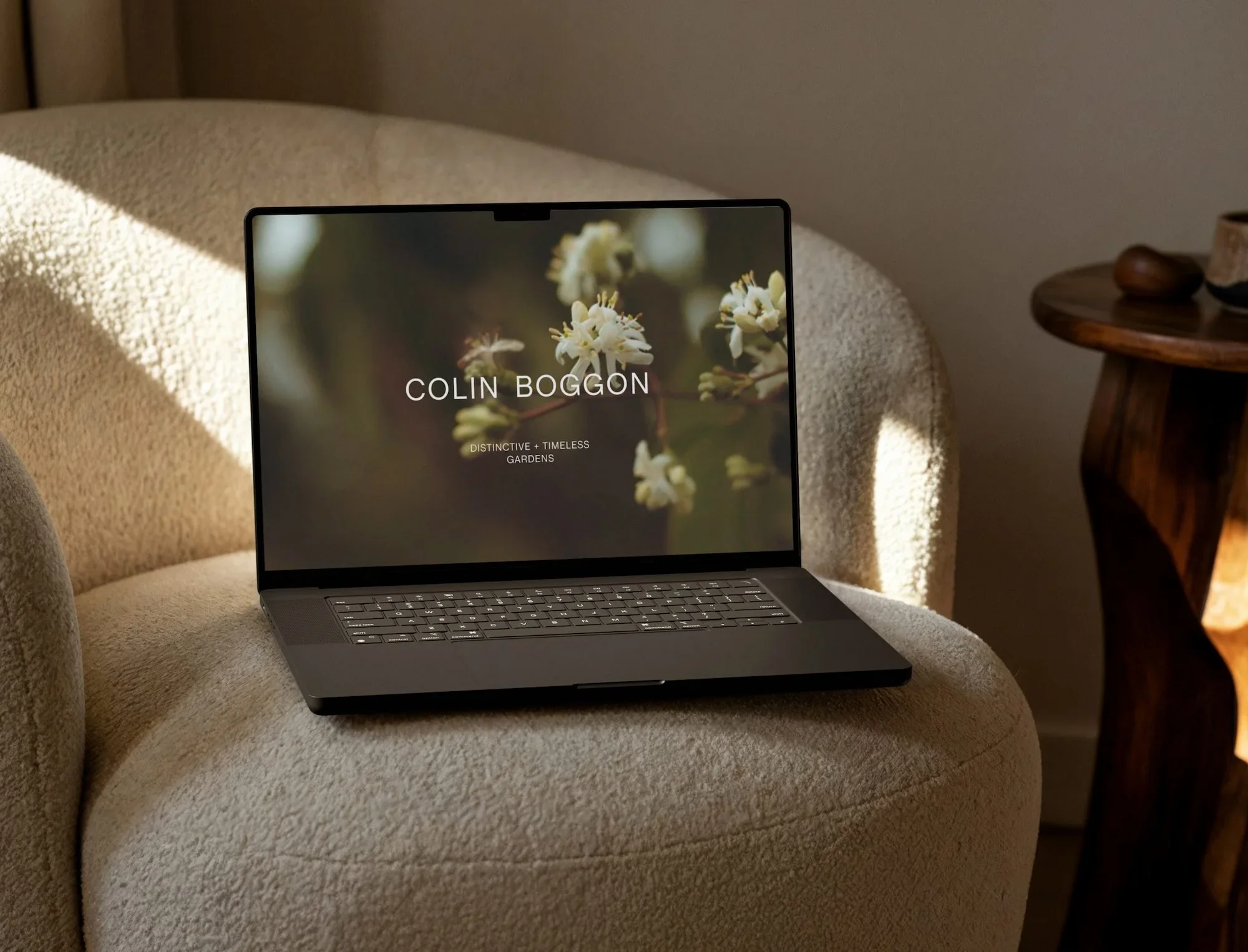 Laptop on a textured chair displaying the Colin Boggon garden design website with a floral background and minimalist text