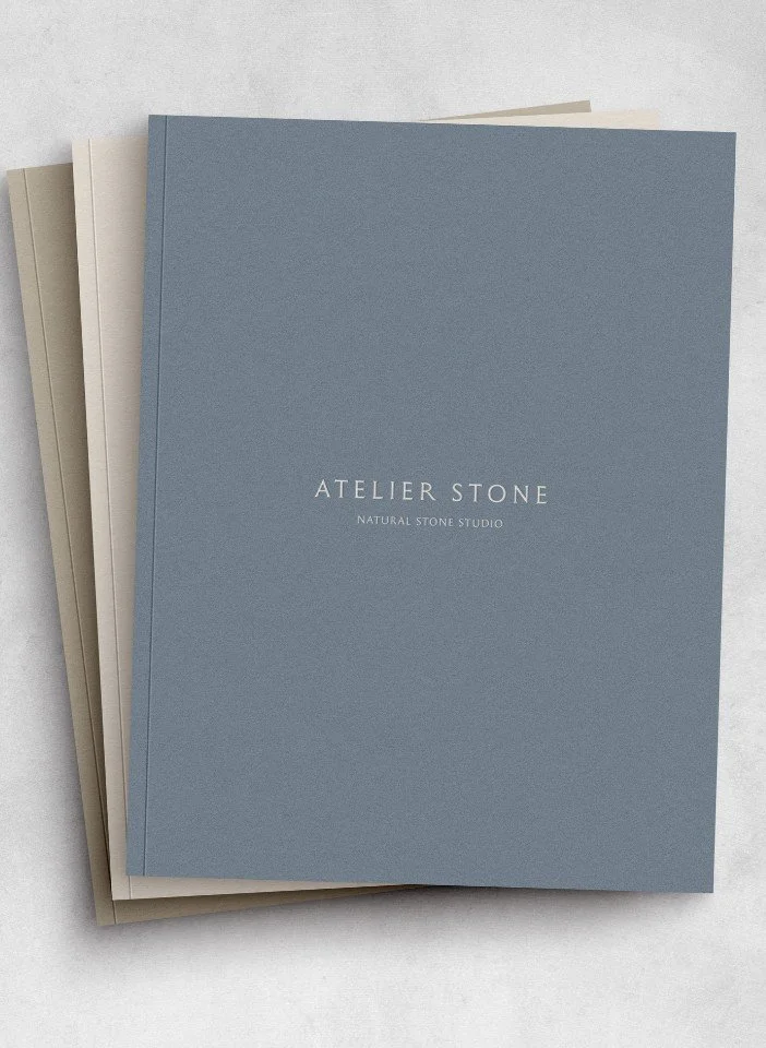 Stack of Atelier Stone Natural Stone Studio catalogues featuring muted blue and beige textured paper covers on grey stone
