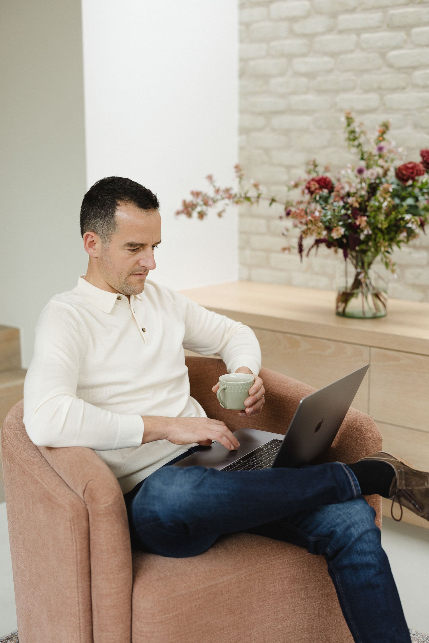 A male designer sitting in a peach armchair working on a laptop while holding a green ceramic mug in a bright modern room