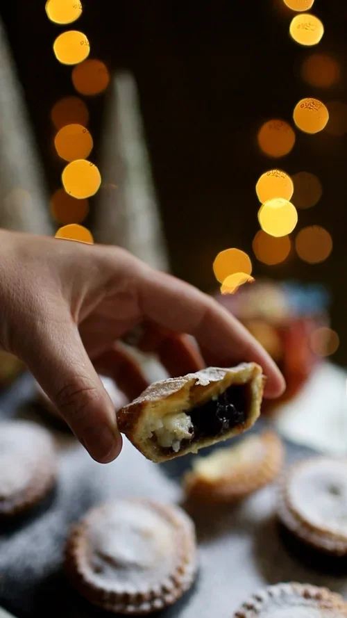 Hand holding a halved mince pie showing a dark fruity filling alongside a white cream cheese layer and a dusted pastry lid