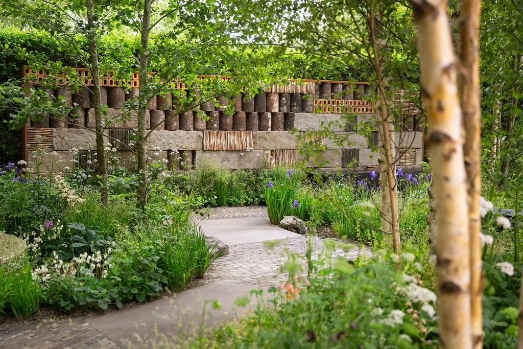 Stone path winding through silver birch trees towards a decorative wall made of stacked clay pipes and textured blocks