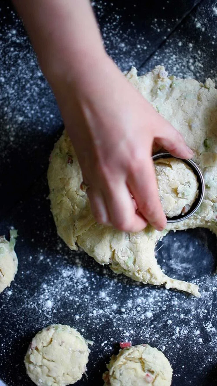 Hand using a metal circular cutter to stamp out round scones from a flat layer of dough on a floured dark surface
