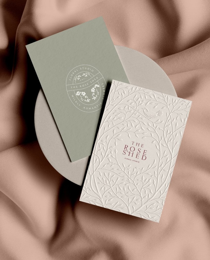 Two branded cards on a pink fabric background including an ivory card with an embossed bird and leaf wreath pattern