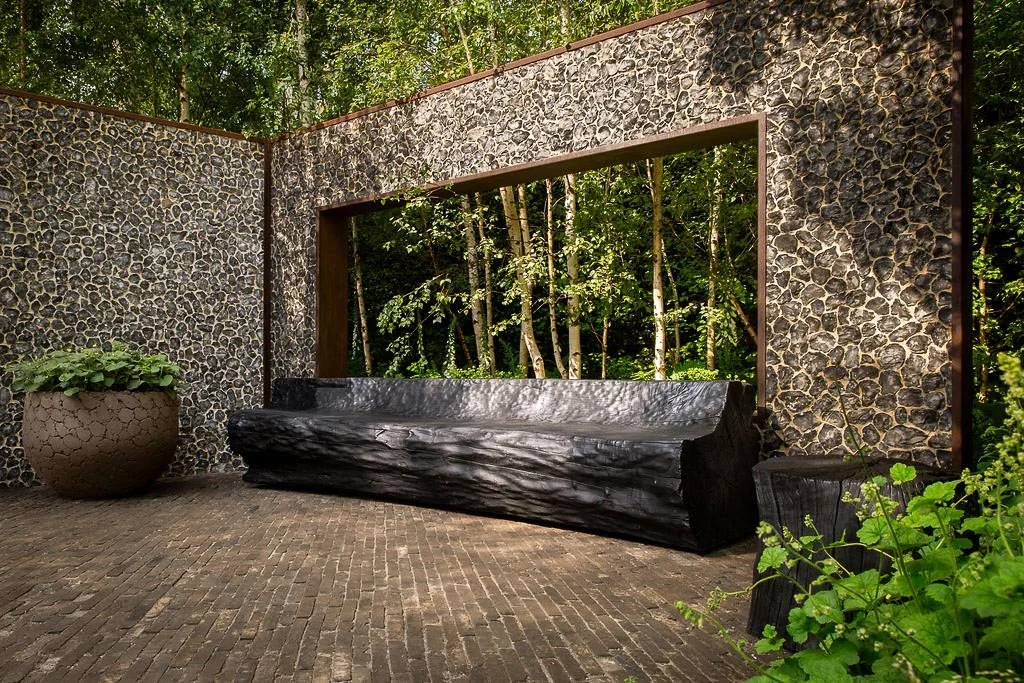 Hand-carved black log bench sits on a brick patio before a flint stone wall with a rectangular cutout showing birch trees