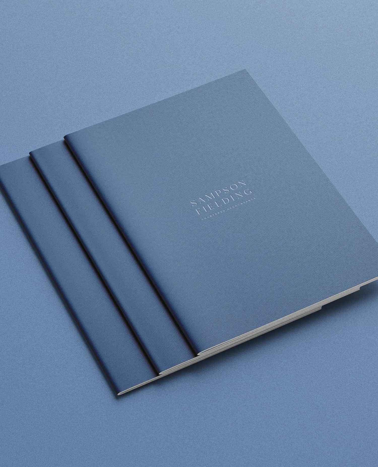 Three navy blue textured notebooks with the Sampson Fielding logo centered in silver foil stacked on a matching blue surface