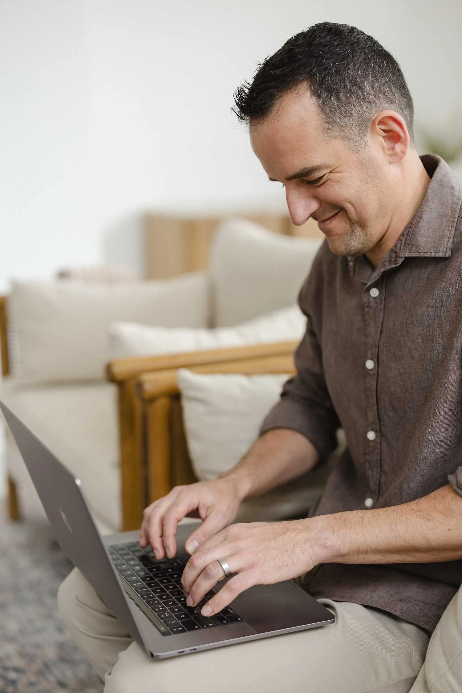 Close up of a smiling man in a brown shirt typing on a laptop while sitting in a bright and airy modern home office space