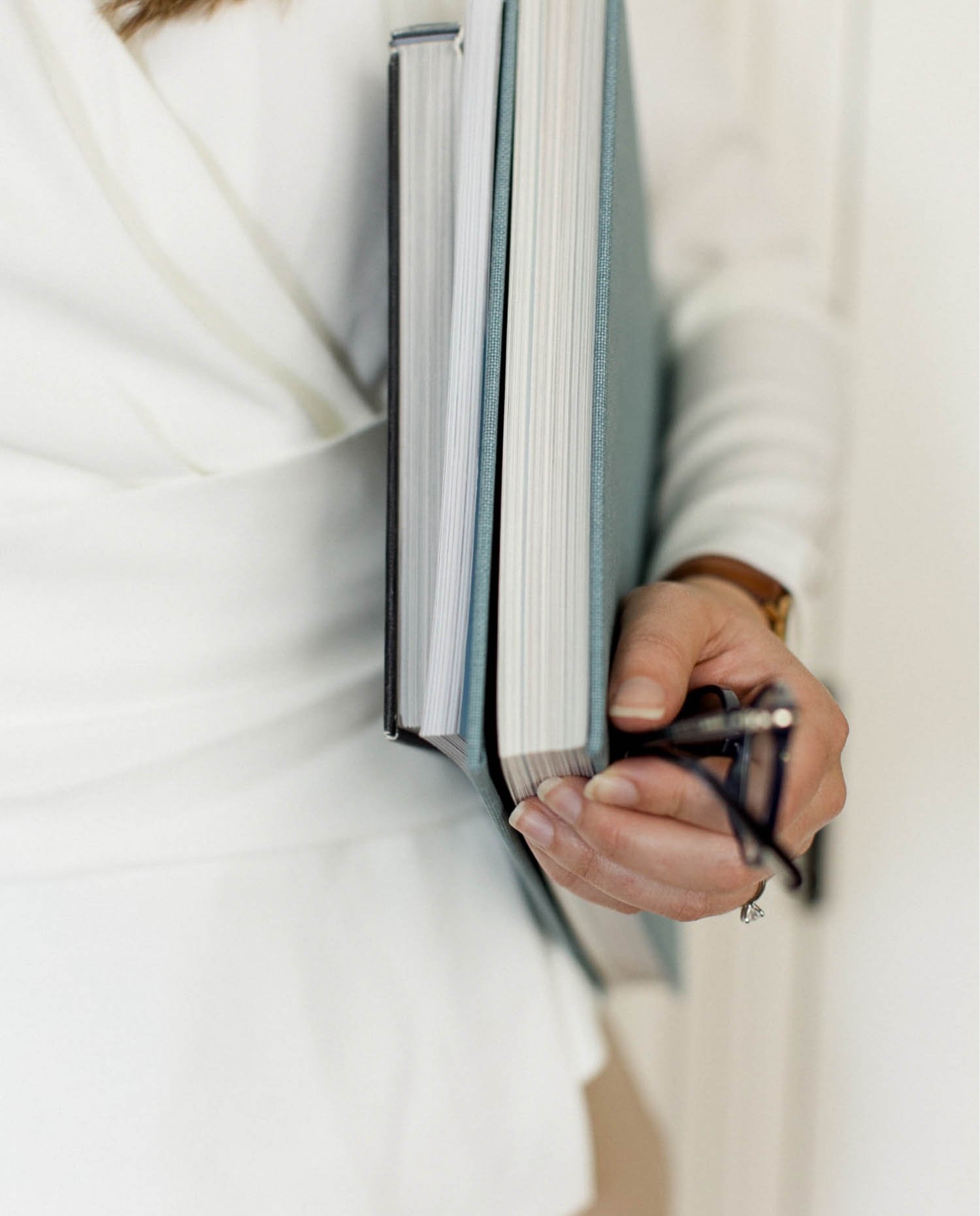 Close up of a person in a white blazer holding three light blue notebooks and a pair of dark framed glasses with one hand