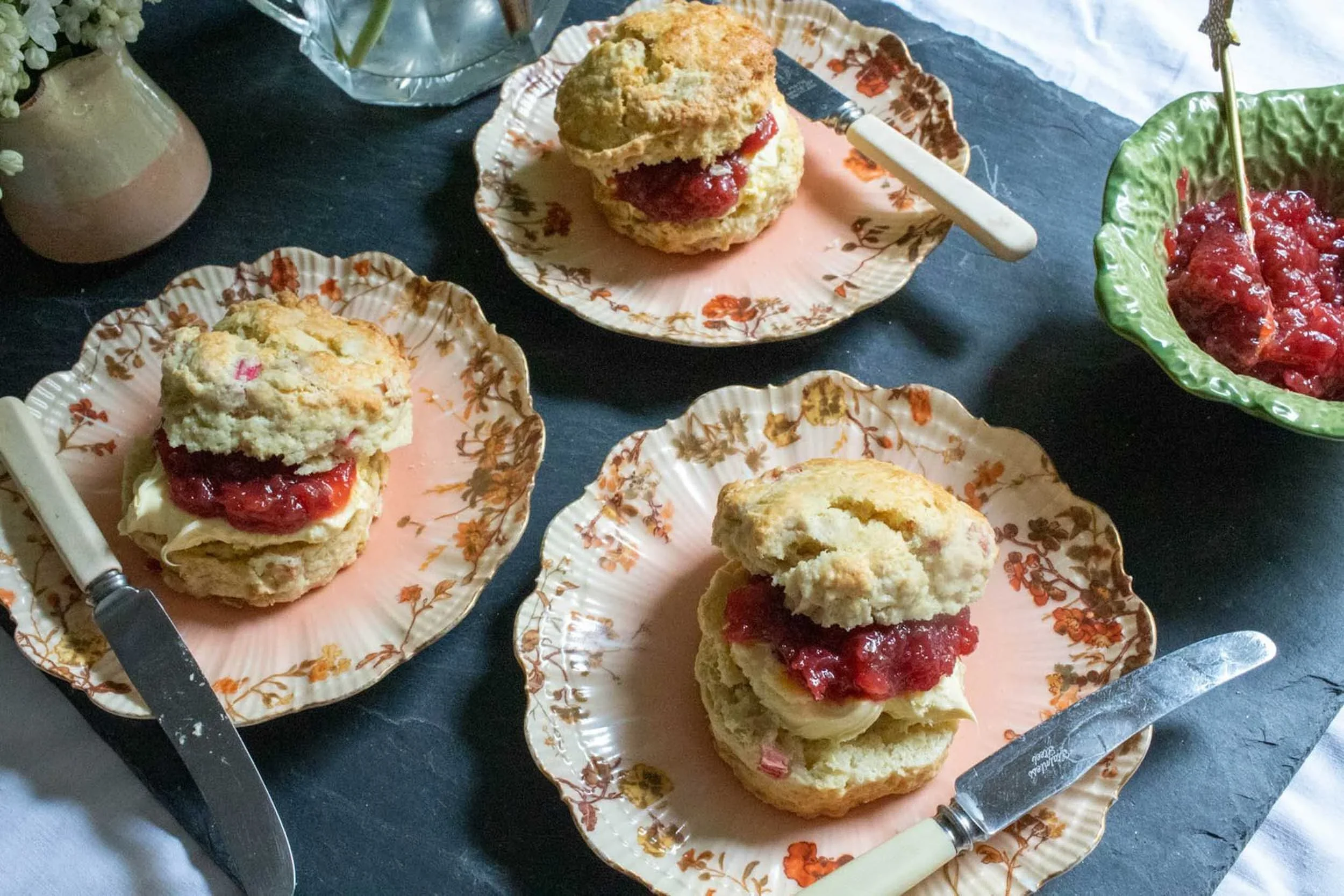 Three rhubarb scones with cream and jam on floral plates served with vintage knives and a bowl of extra fruit preserve