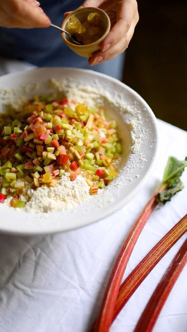 Hand adding chopped crystallised ginger from a small bowl onto a pile of diced rhubarb and flour in a large bowl