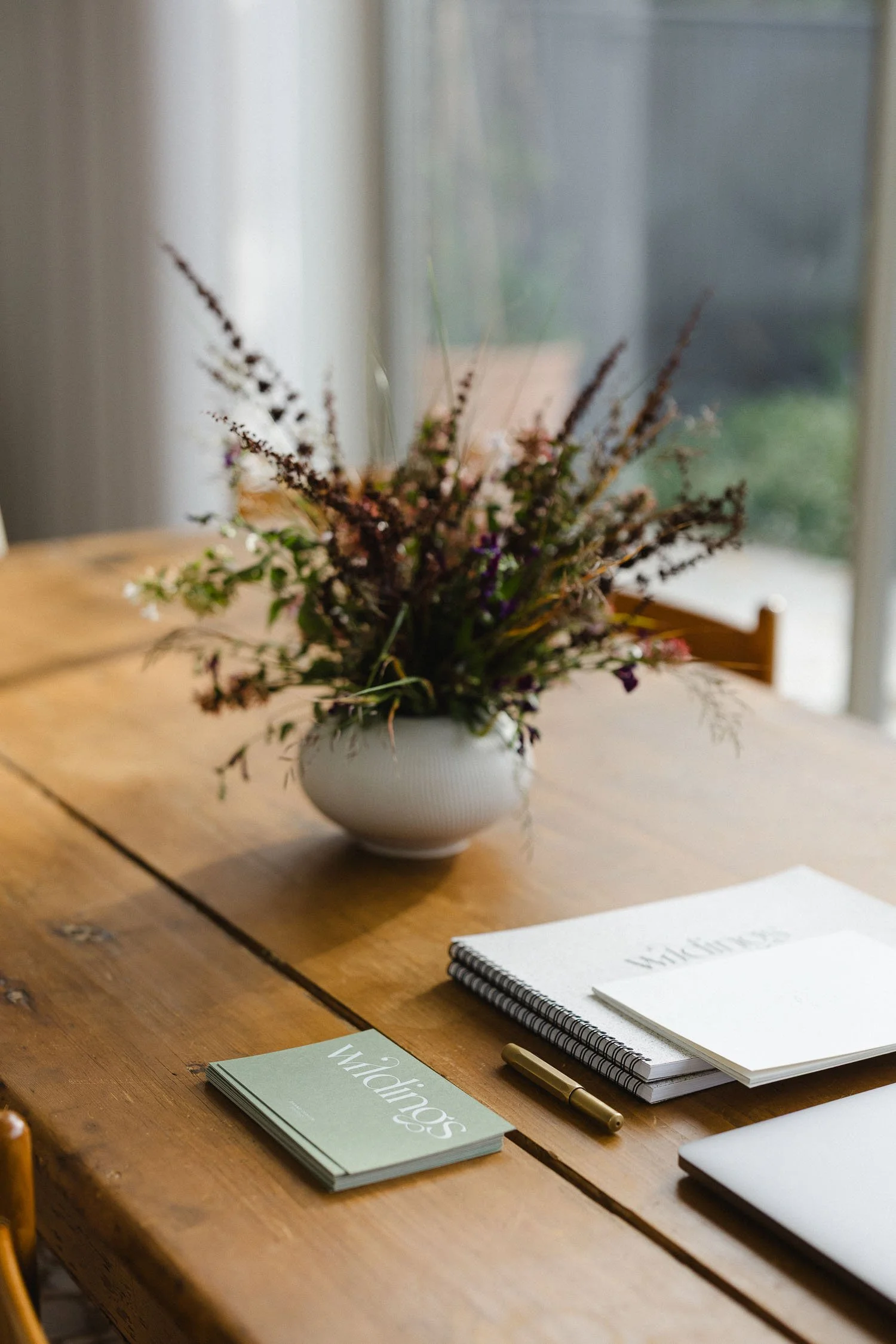 Branded Wildings notebooks and business cards on a wooden desk next to a white vase filled with dried wildflowers and a pen