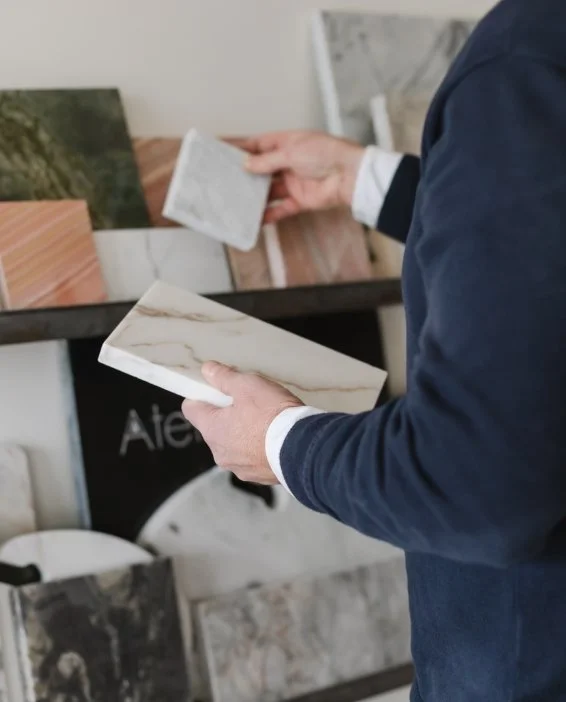A person in a navy jacket holding a white marble sample while browsing various stone slabs on a display shelf