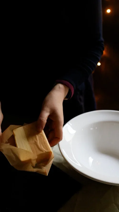 Person holding a block of butter wrapped in brown greaseproof paper over a white mixing bowl in a dimly lit kitchen