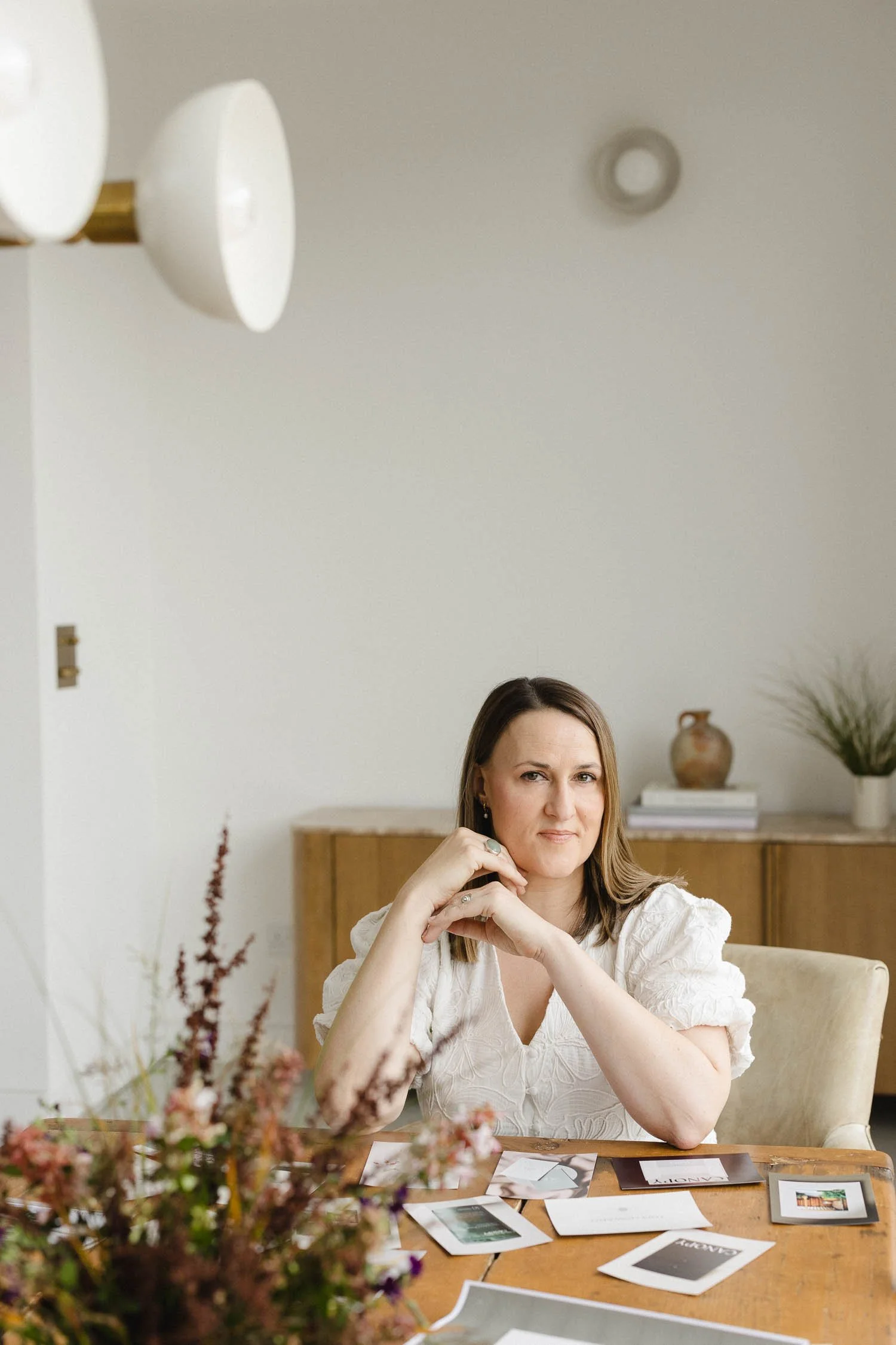 Professional headshot of a woman in a white blouse sitting in a bright studio with neutral wooden furniture