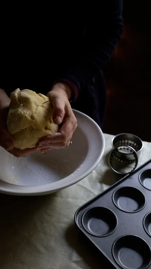 Hands kneading a ball of smooth yellow pastry dough over a white ceramic bowl next to a metal baking tray and pastry cutters