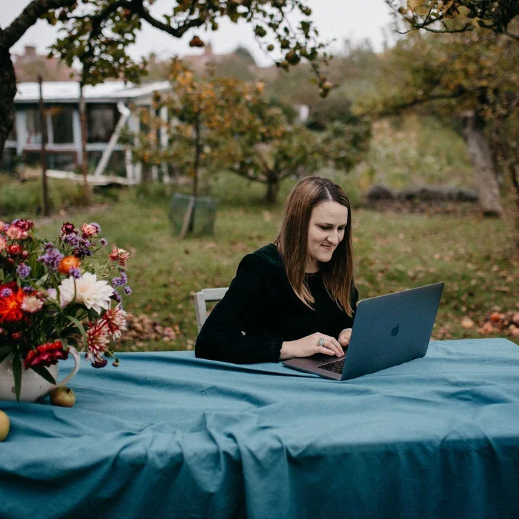 A woman sits at a garden table working on a laptop next to a vibrant bouquet of flowers in a white ceramic jug