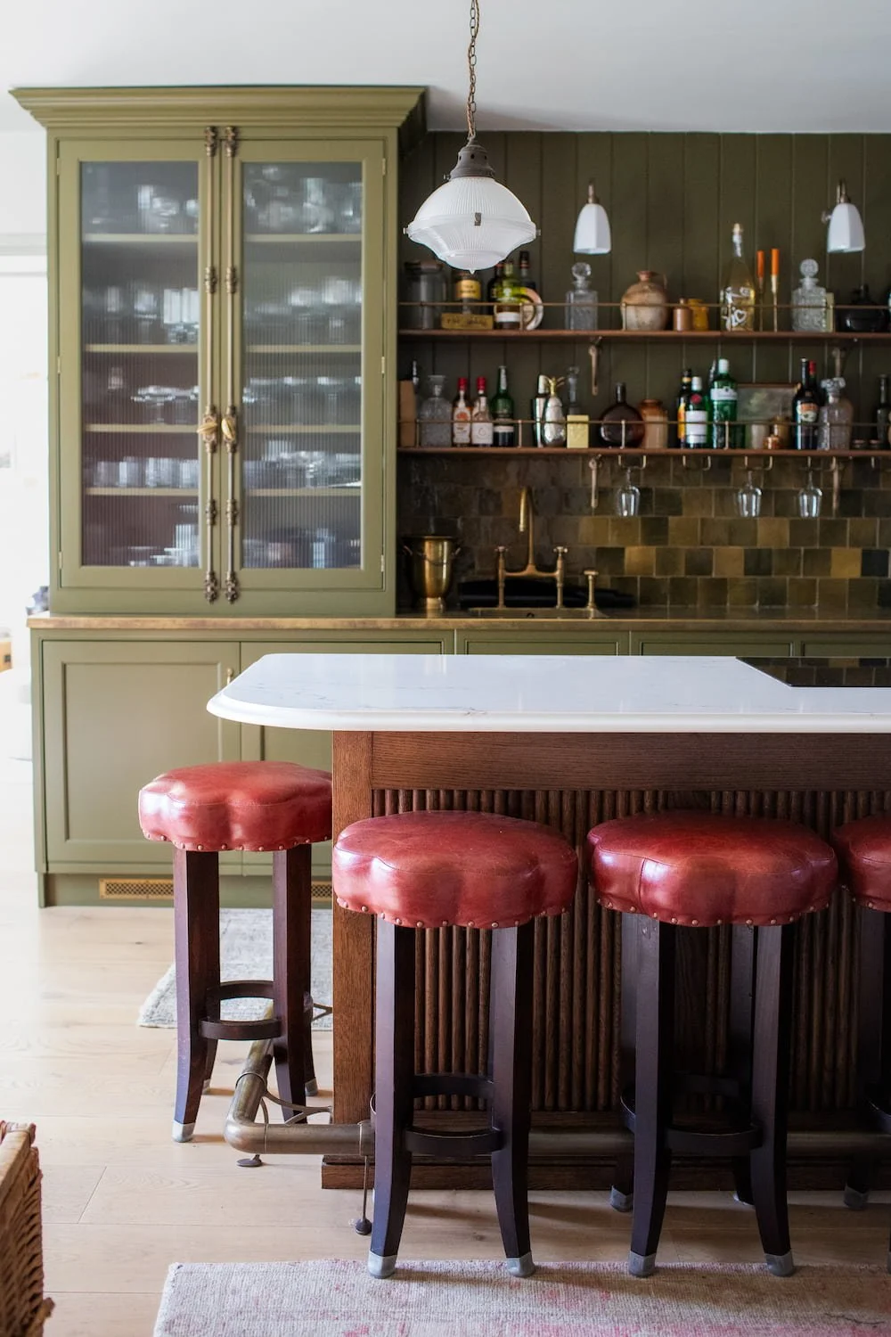 Kitchen island with a white marble top and dark wood base featuring four round red leather stools with brass stud details