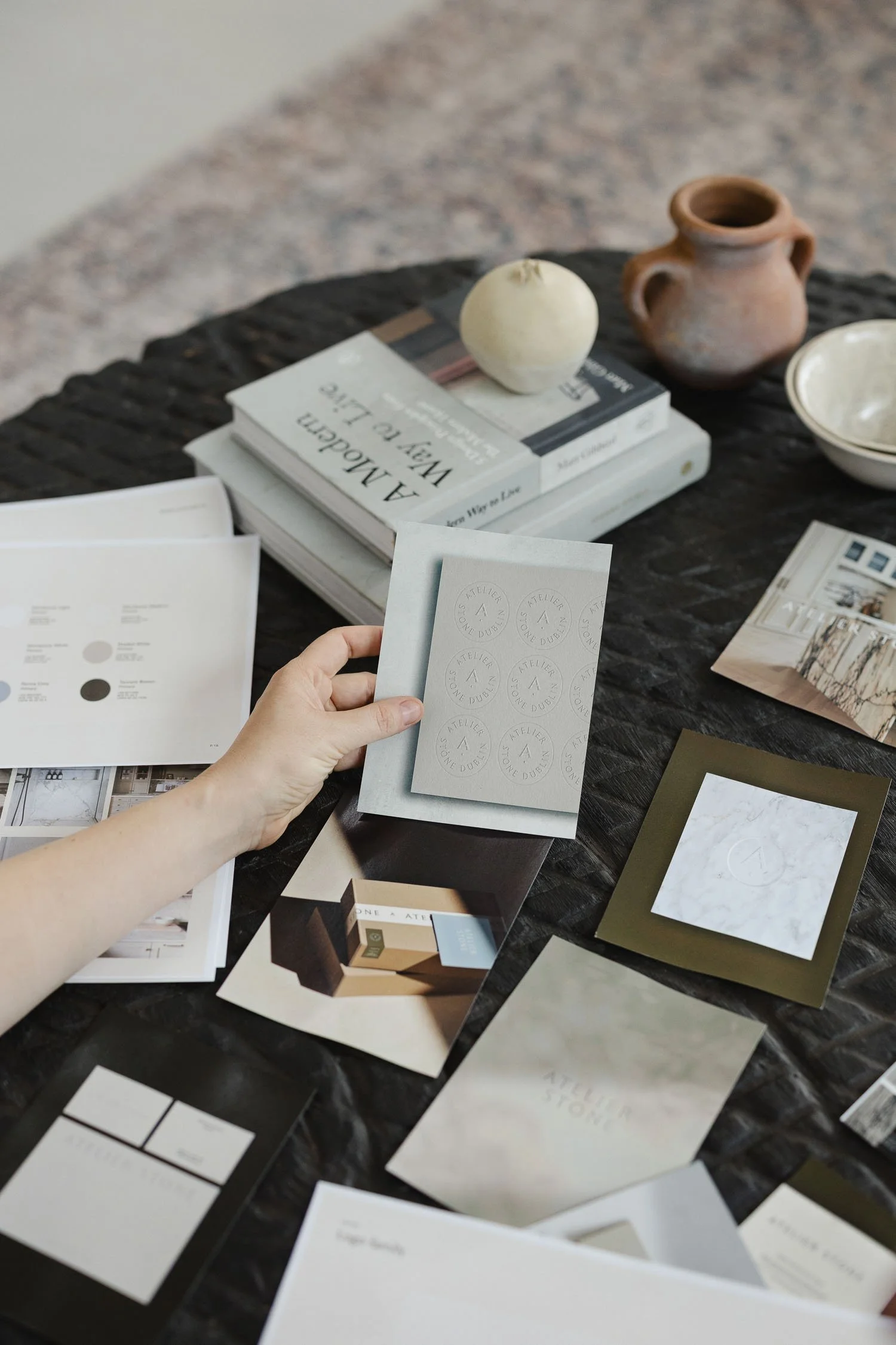 Person holding an embossed logo card over a table filled with brand identity materials, books and terracotta pottery