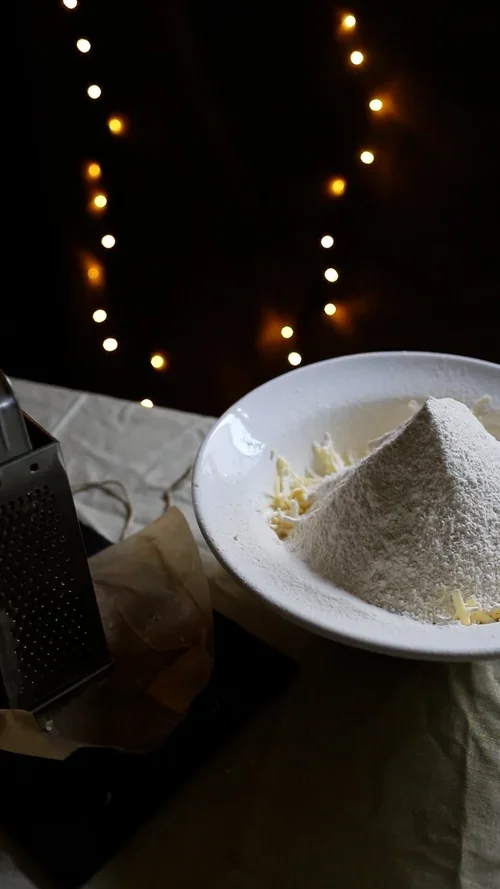 A mound of white flour sits on top of grated butter in a white bowl next to a metal grater and festive fairy lights