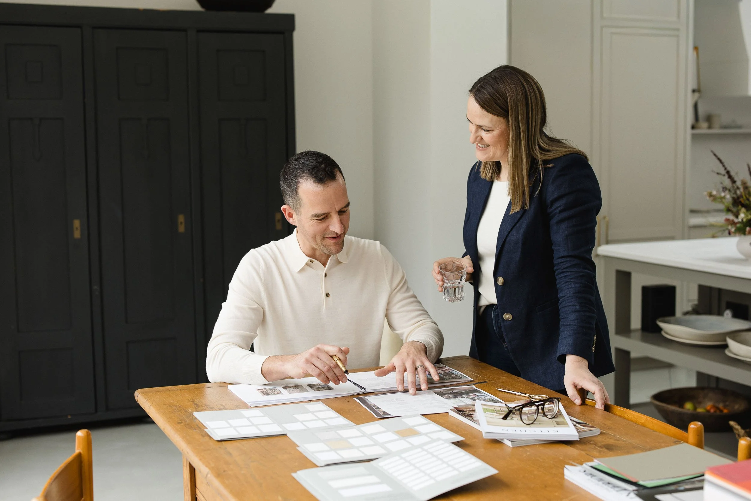 Two designers standing over a wooden table discussing printed interior project plans and material samples in a bright studio
