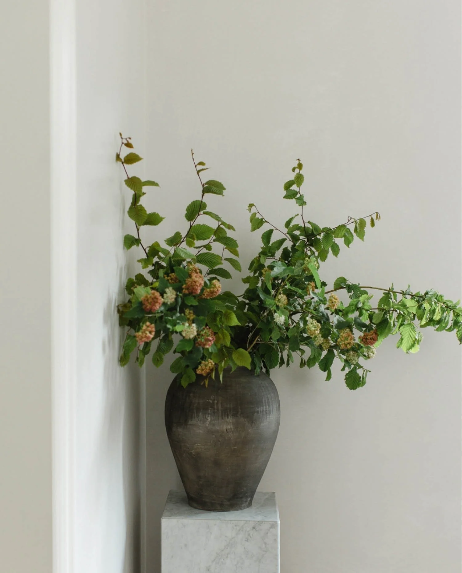 A large dark rustic ceramic vase filled with green leafy branches and small flowers on a marble plinth against a white wall