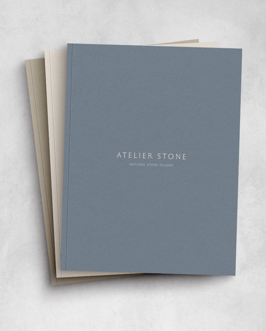 A stack of three notebooks in blue and cream featuring the Atelier Stone Natural Stone Studio logo on a grey background