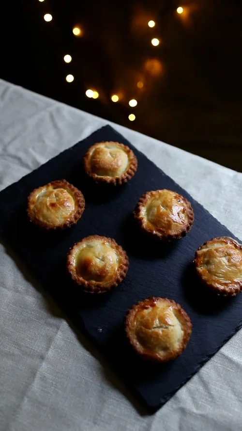 Six golden brown baked mince pies with fluted edges arranged in two rows on a dark slate board over a light linen tablecloth