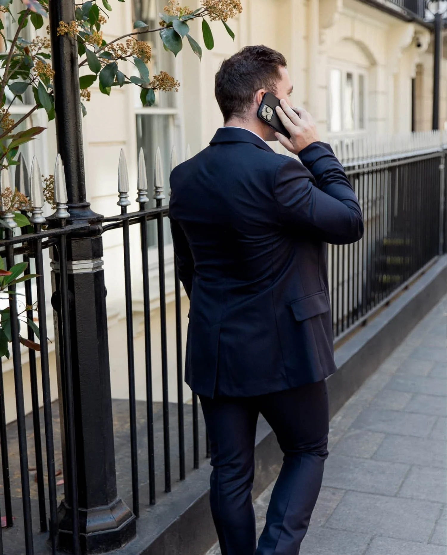 Rear view of a man in a navy suit talking on a mobile phone while walking past a traditional Mayfair building with railings