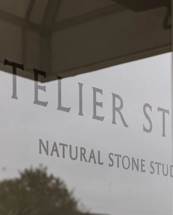 Close up of the Atelier Stone Natural Stone Studio logo etched in serif font on a glass window reflecting the outdoors