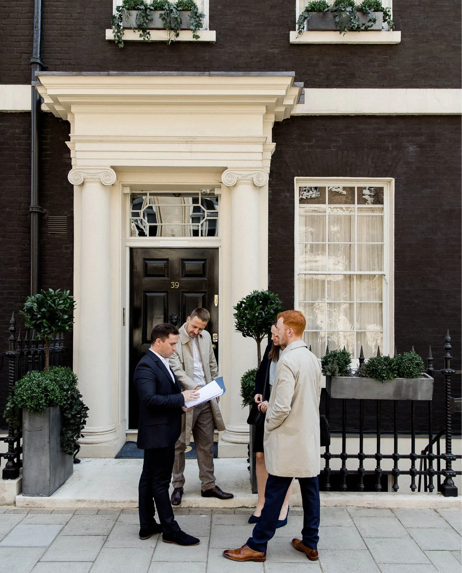 Four professionals in suits and coats stand outside a black Mayfair door at number 39 reviewing a paper document together