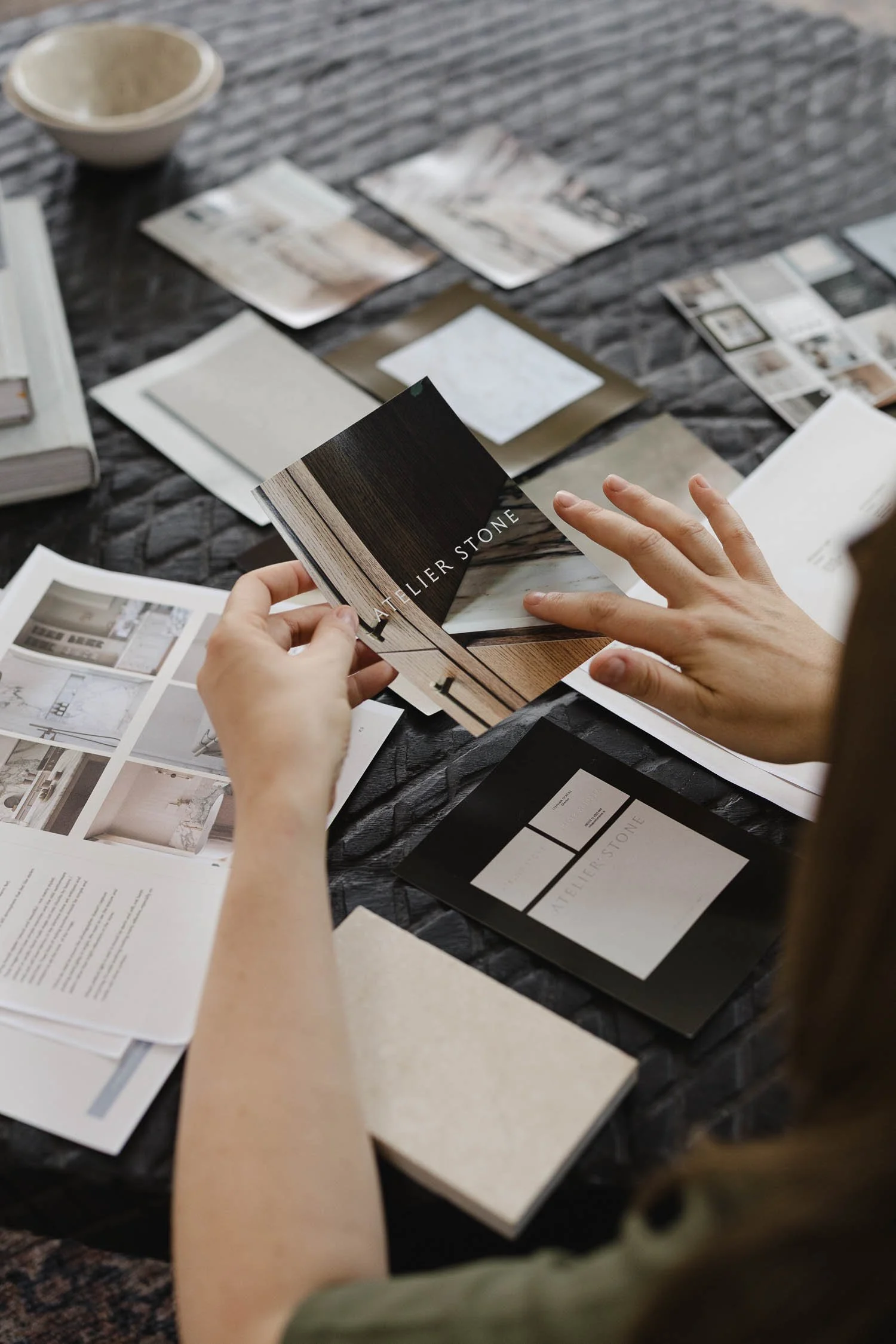Designer holding Atelier Stone branding mockups on a dark textured table with mood board photos and material swatches