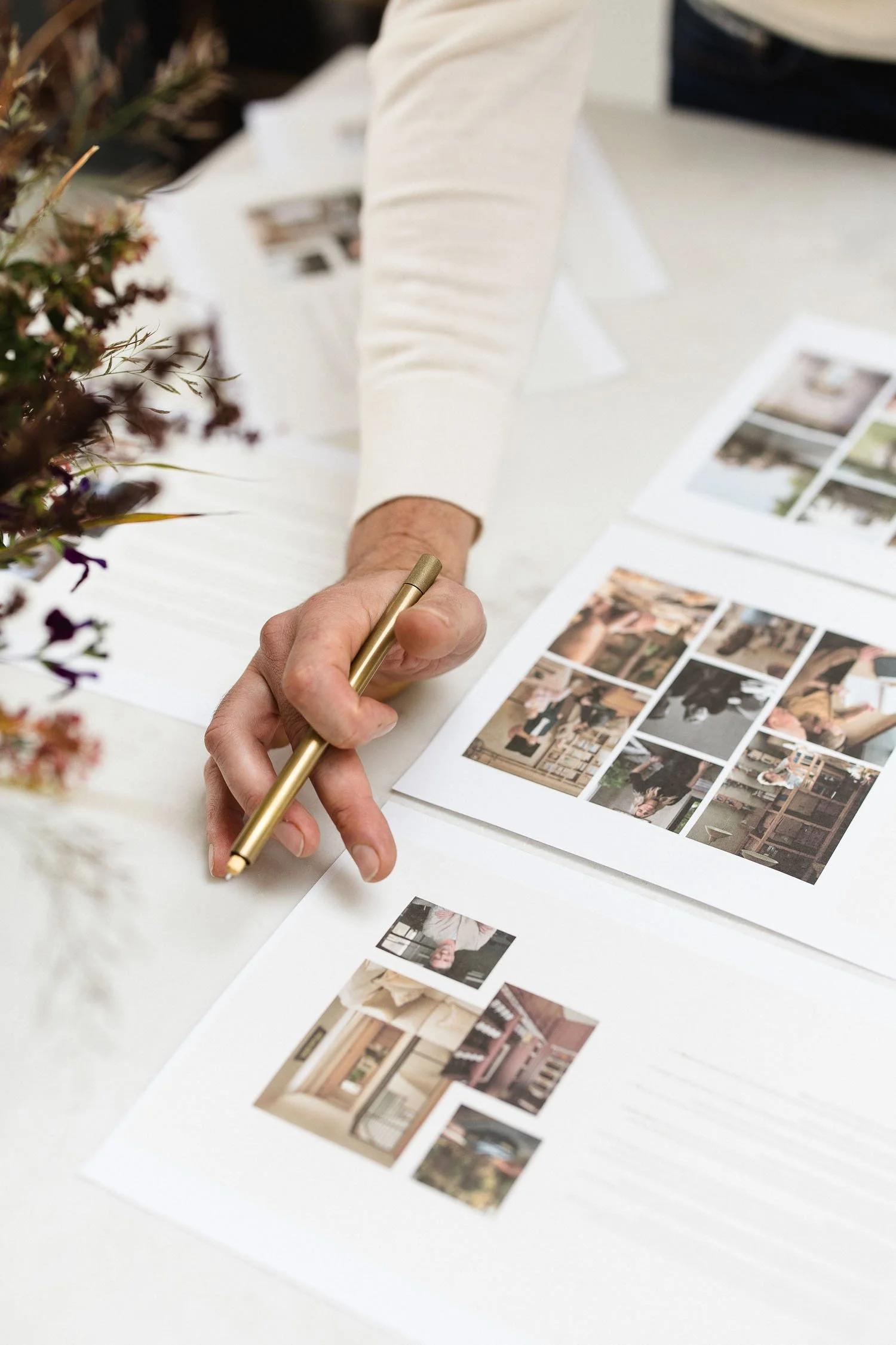 A person using a brass pen to point at interior design mood board sheets with various room photos on a white desk