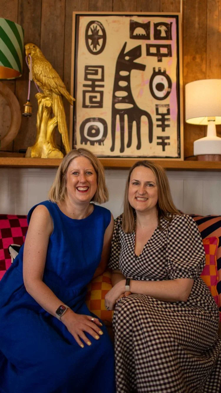Two women smiling at a business social event sitting on a sofa in front of a golden parrot statue and modern framed art