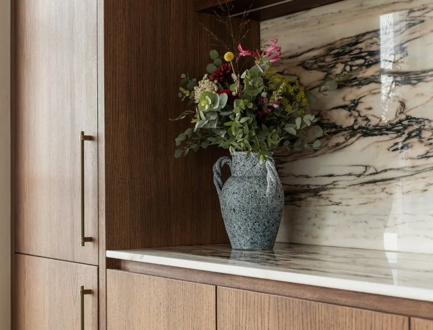 A textured stone vase with flowers on a white marble worktop against a dark marble backsplash and natural oak cabinetry